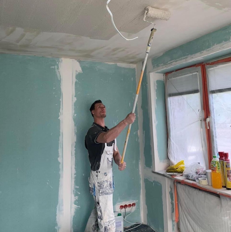 Mężczyzna w białym kombinezonie maluje sufit wałkiem na teleskopowym kiju w pomieszczeniu z płytami kartonowo-gipsowymi, widoczne puszki z farbą przy oknie.