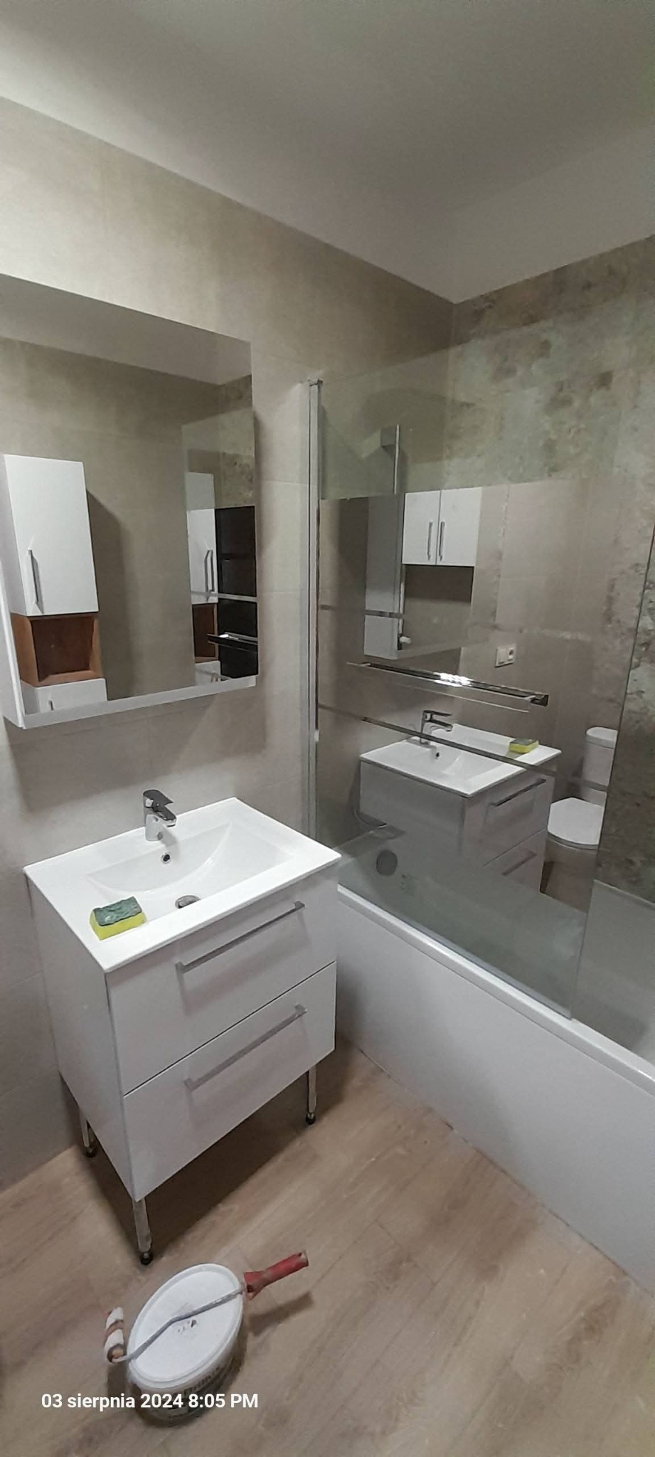 Nowoczesna łazienka z umywalką na szafce, lustrem z szafką i wanną za szklanym parawanem. Na podłodze leży wałek malarski.