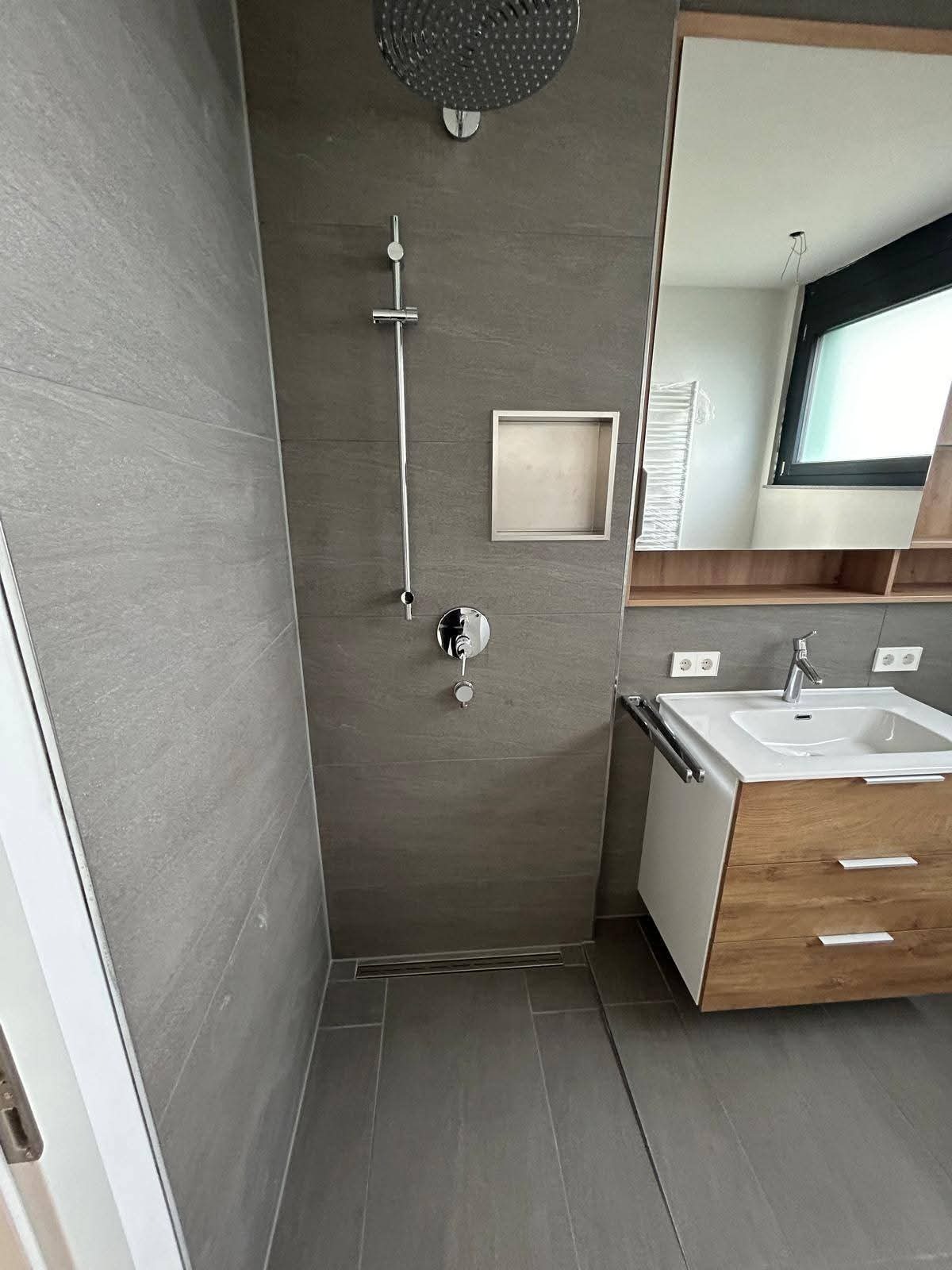 Nowoczesna łazienka z szarymi płytkami, deszczownicą, wnęką i drewnianą szafką z umywalką. Minimalistyczny design i stonowana kolorystyka.