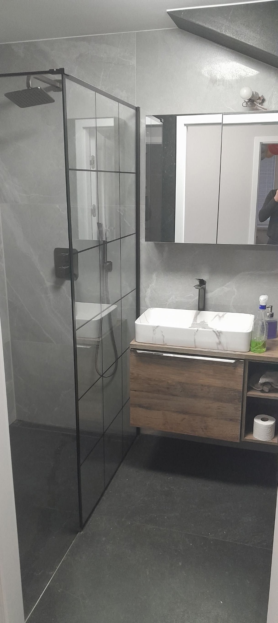 Nowoczesna łazienka z kabiną typu walk-in, marmurową umywalką i drewnianą szafką. Szare płytki imitujące marmur na ścianach i podłodze. Minimalistyczny design.