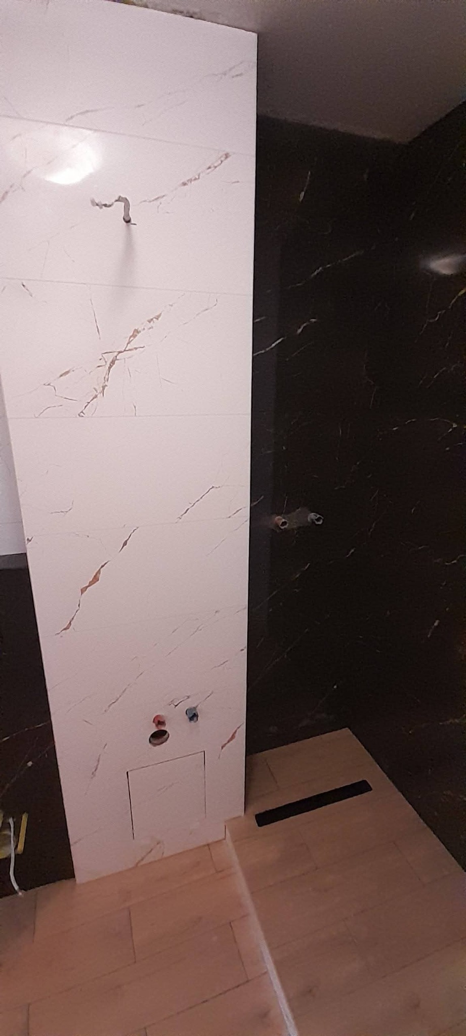 Wykończona ściana w łazience z białych i czarnych płytek imitujących marmur, z widocznymi przyłączami wodnymi i odpływem liniowym w podłodze.
