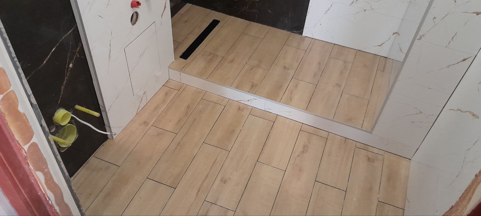 Wykończona kabina prysznicowa z drewnopodobnymi płytkami na podłodze i marmurowymi na ścianach. Widoczny czarny odpływ liniowy i fragment instalacji.