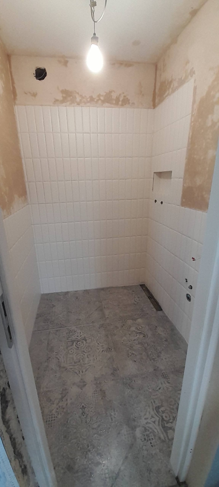 Wykończona łazienka z białymi, prostokątnymi płytkami na ścianach i dekoracyjną szarą podłogą. Widoczny otwór na półkę w ścianie i niedokończone wykończenie.