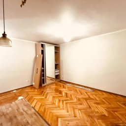 Artur Solo - Pusta, jasna przestrzeń z parkietem w jodełkę, częściowo zabezpieczonym folią malarską, z rozłożonymi elementami szafy przesuwnej IKEA i wiszącą lampą retro.