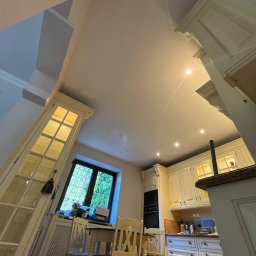 Artur Solo - Ujęcie z dołu na jasną kuchnię w domu z wysokim sufitem, widoczne białe meble, okno z widokiem na zieleń i część jadalni z krzesłami.