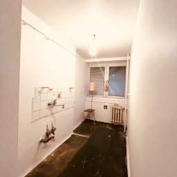 Artur Solo - Wnętrze małej kuchni w trakcie remontu: ściany pomalowane na biało, częściowo usunięte płytki nad zlewem, odsłonięte rury, stara podłoga z resztkami wykładziny, grzejnik pod oknem, lampka na stoliku.