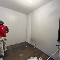 Artur Solo - Malarz w czerwonej koszulce maluje ścianę na biało za pomocą wałka na długim kiju; widoczne zabezpieczenia taśmą malarską przy podłodze i futrynie drzwiowej.