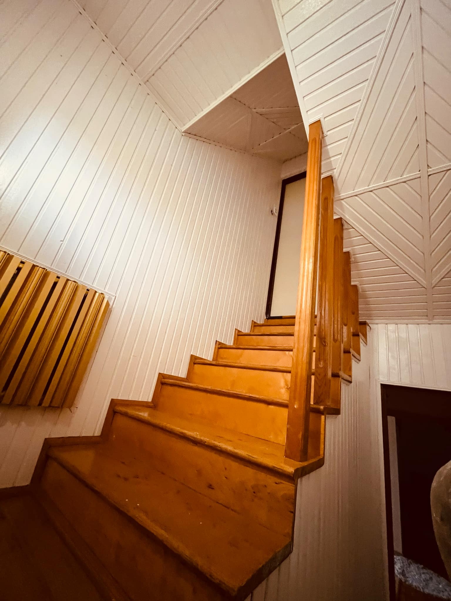 Drewniane schody z balustradą, prowadzące na górę, otoczone białymi, panelowymi ścianami i sufitem, widoczne z dołu.