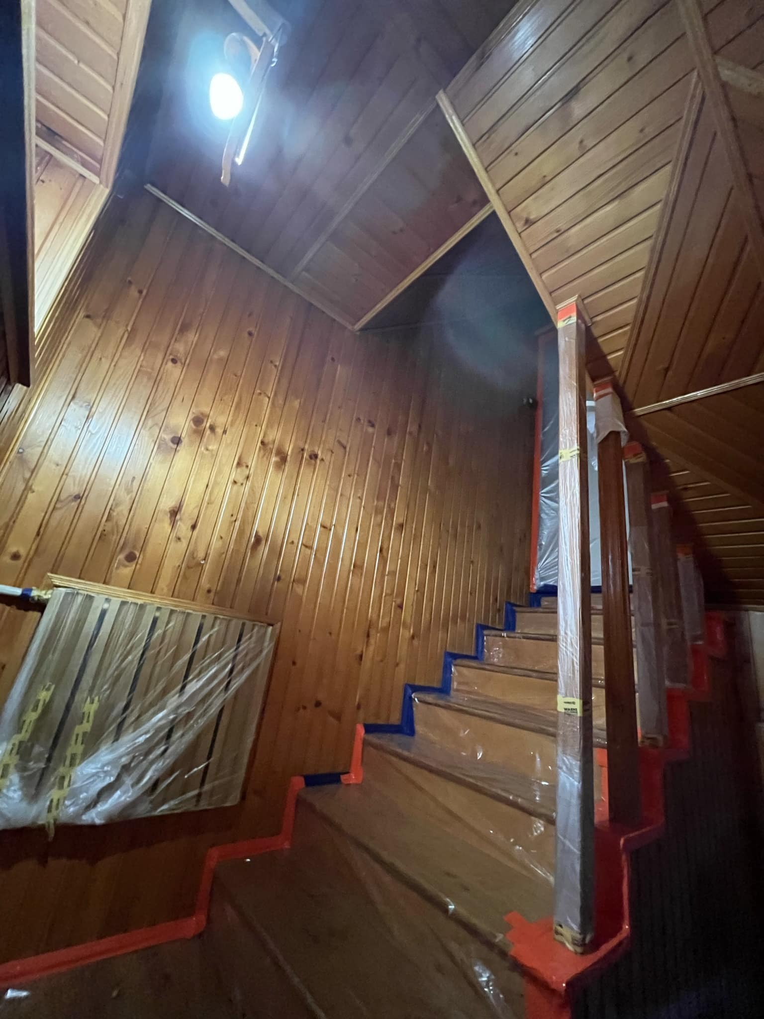 Klatka schodowa w drewnianym domu zabezpieczona folią i taśmą malarską przed malowaniem ścian i schodów, widok z dołu.