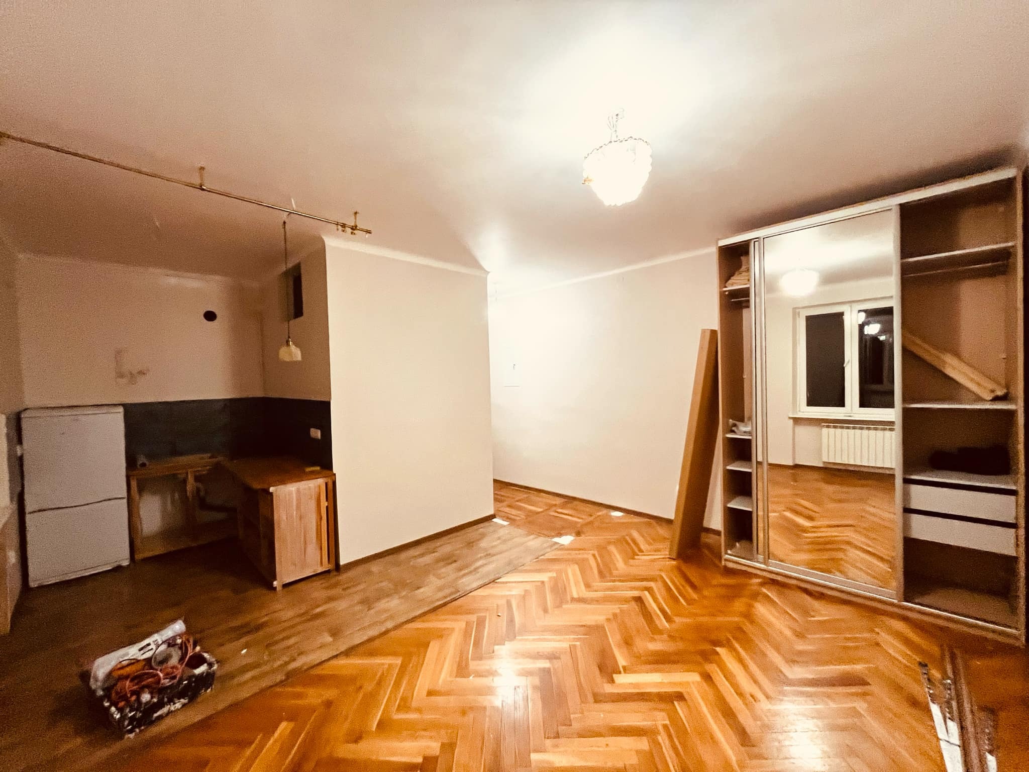 Pokój w trakcie remontu z parkietem w jodełkę, częściowo zakrytym, widoczne narzędzia w koszu, odsłonięta kuchnia z ciemnymi płytkami i szafkami bez frontów, przesuwana szafa z lustrami.