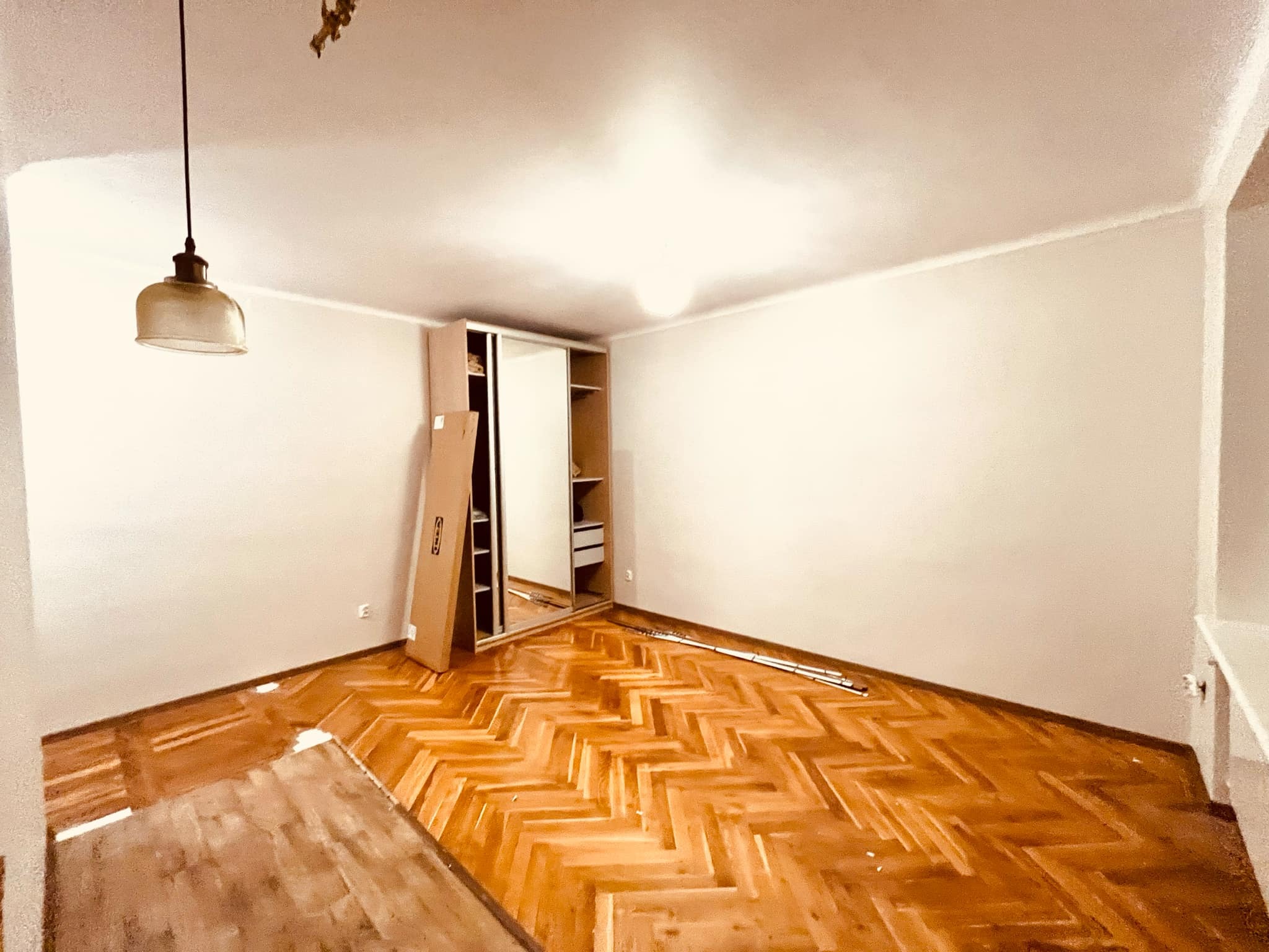 Pusta, jasna przestrzeń z parkietem w jodełkę, częściowo zabezpieczonym folią malarską, z rozłożonymi elementami szafy przesuwnej IKEA i wiszącą lampą retro.