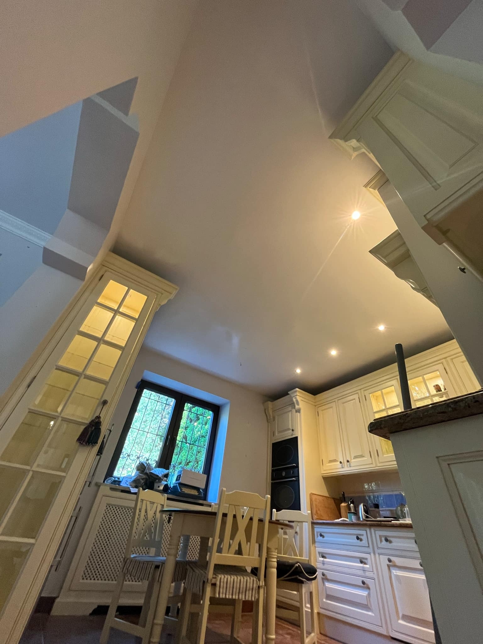 Ujęcie z dołu na jasną kuchnię w domu z wysokim sufitem, widoczne białe meble, okno z widokiem na zieleń i część jadalni z krzesłami.