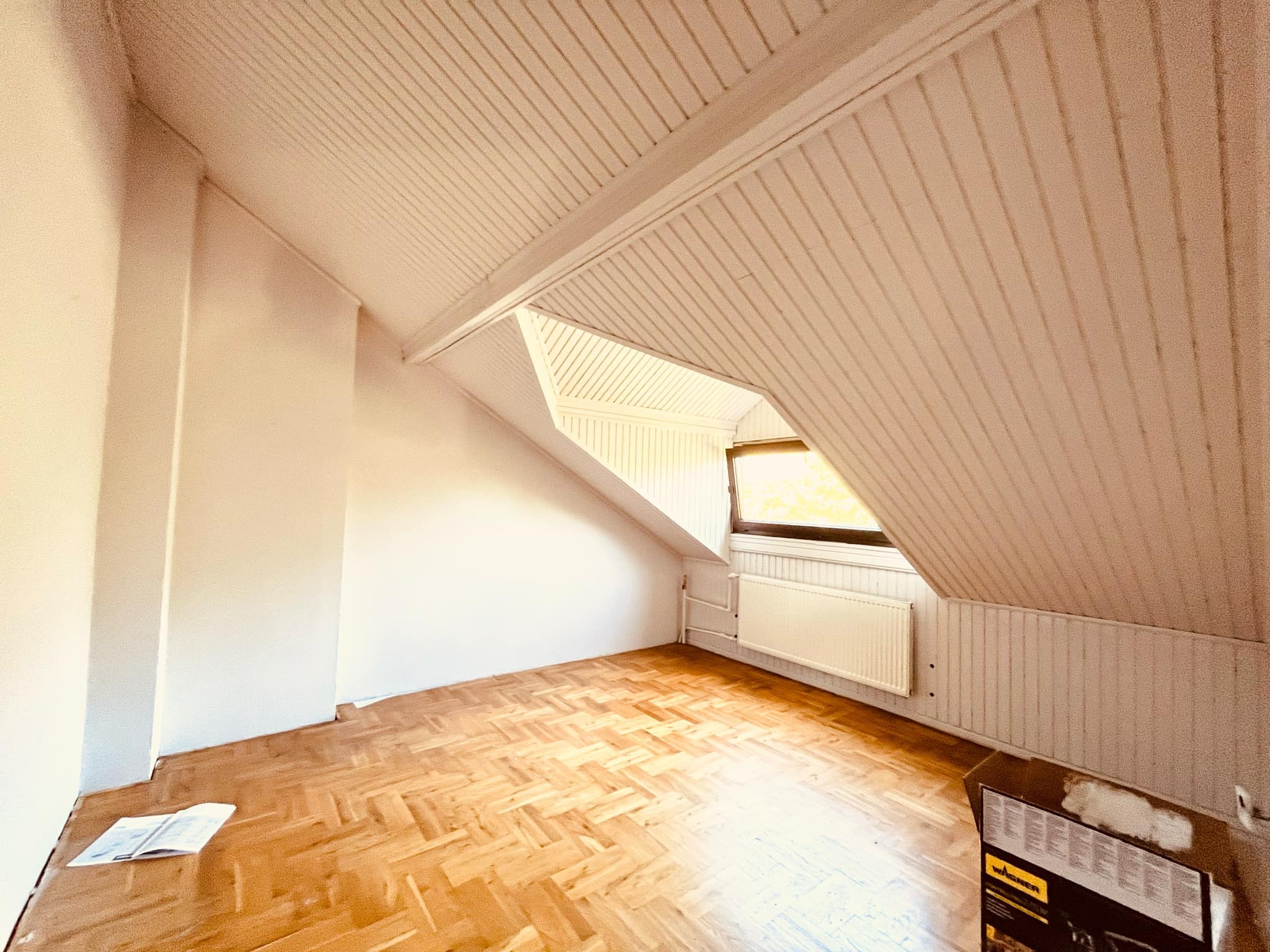 Poddasze z drewnianą boazerią i parkietem przed malowaniem, widoczny grzejnik i okno dachowe, karton po urządzeniu malarskim w rogu.