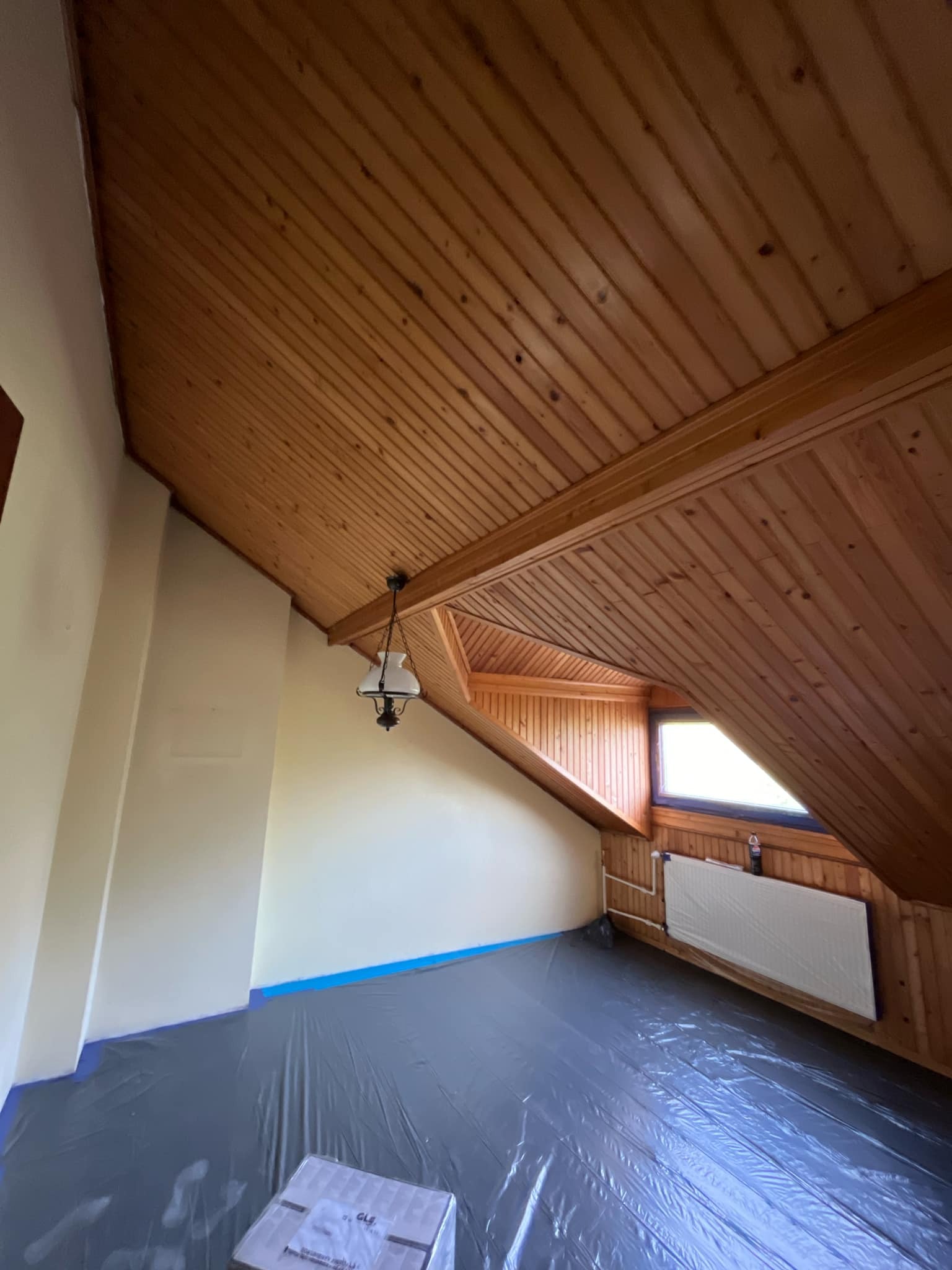 Poddasze zabezpieczone folią malarską przed malowaniem ścian, widoczny grzejnik i drewniana boazeria na suficie oraz okno dachowe.