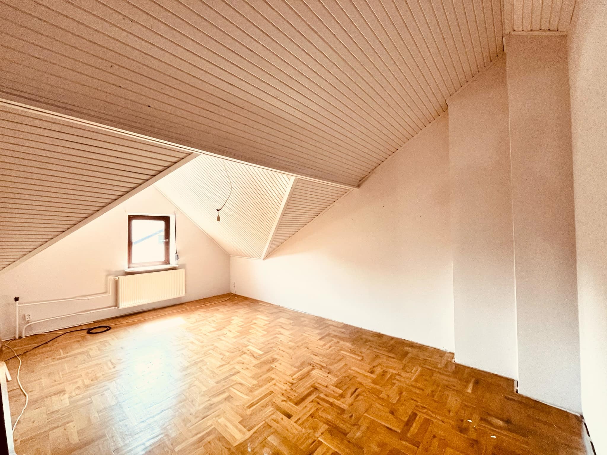 Poddasze z drewnianą boazerią na suficie i parkietem w jodełkę, ściany świeżo pomalowane na biało, widoczny grzejnik pod oknem.