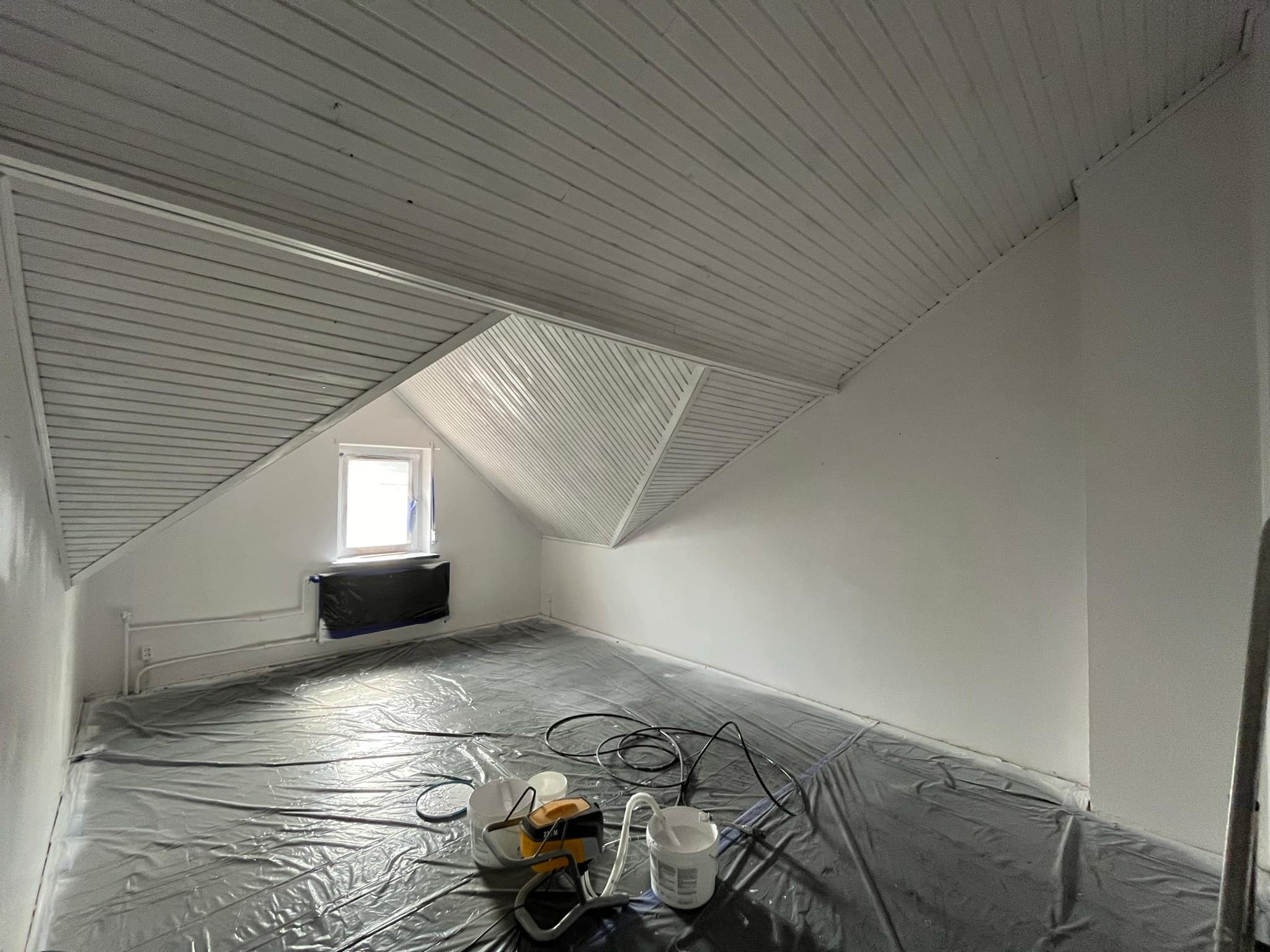 Pokój na poddaszu przygotowany do malowania: zabezpieczona folią podłoga, zakryte okno, widoczny agregat malarski i świeżo pomalowane ściany.