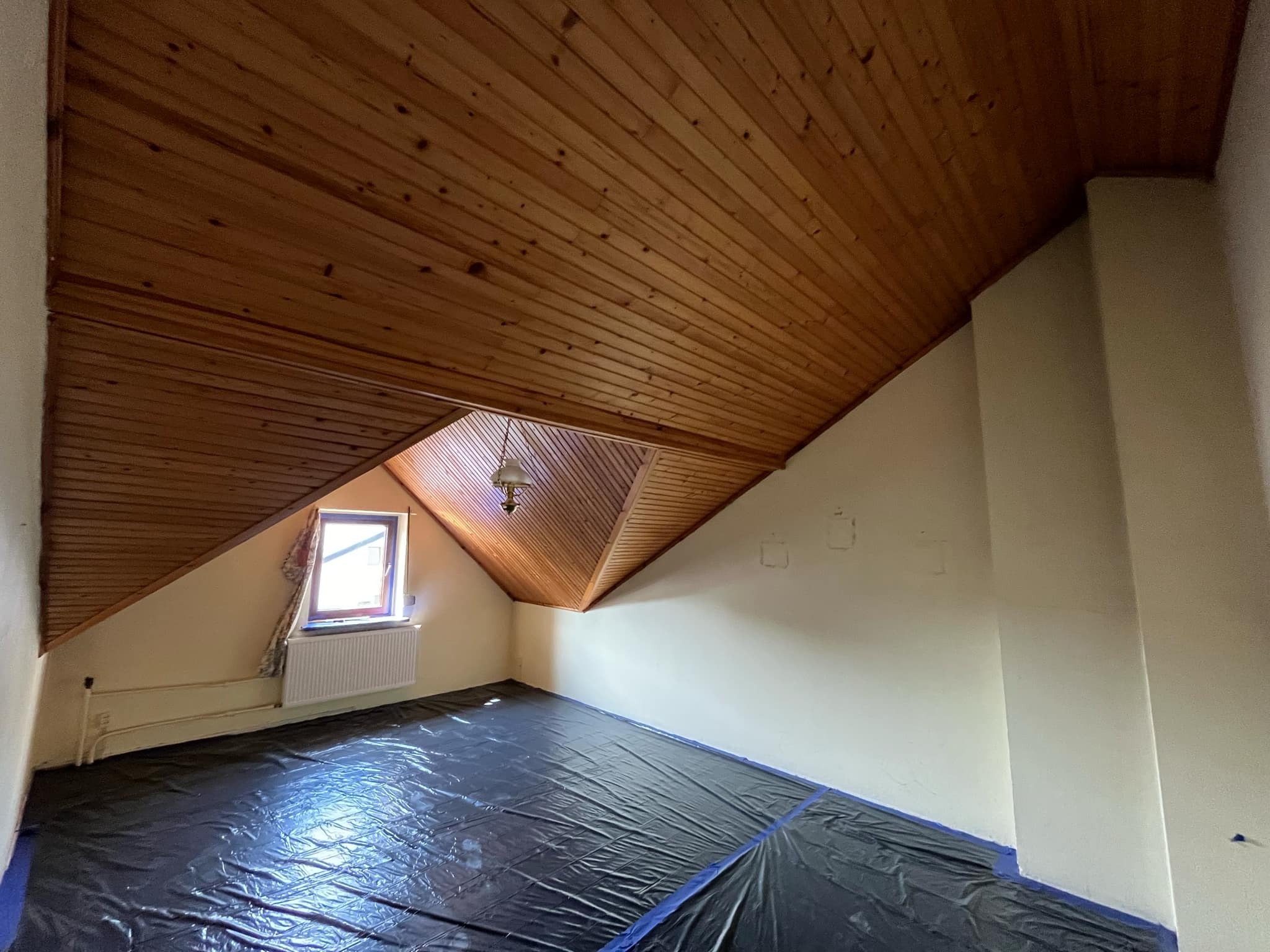 Poddasze zabezpieczone folią malarską przed malowaniem ścian, widoczny skośny drewniany sufit, okno z grzejnikiem i stara lampa sufitowa.