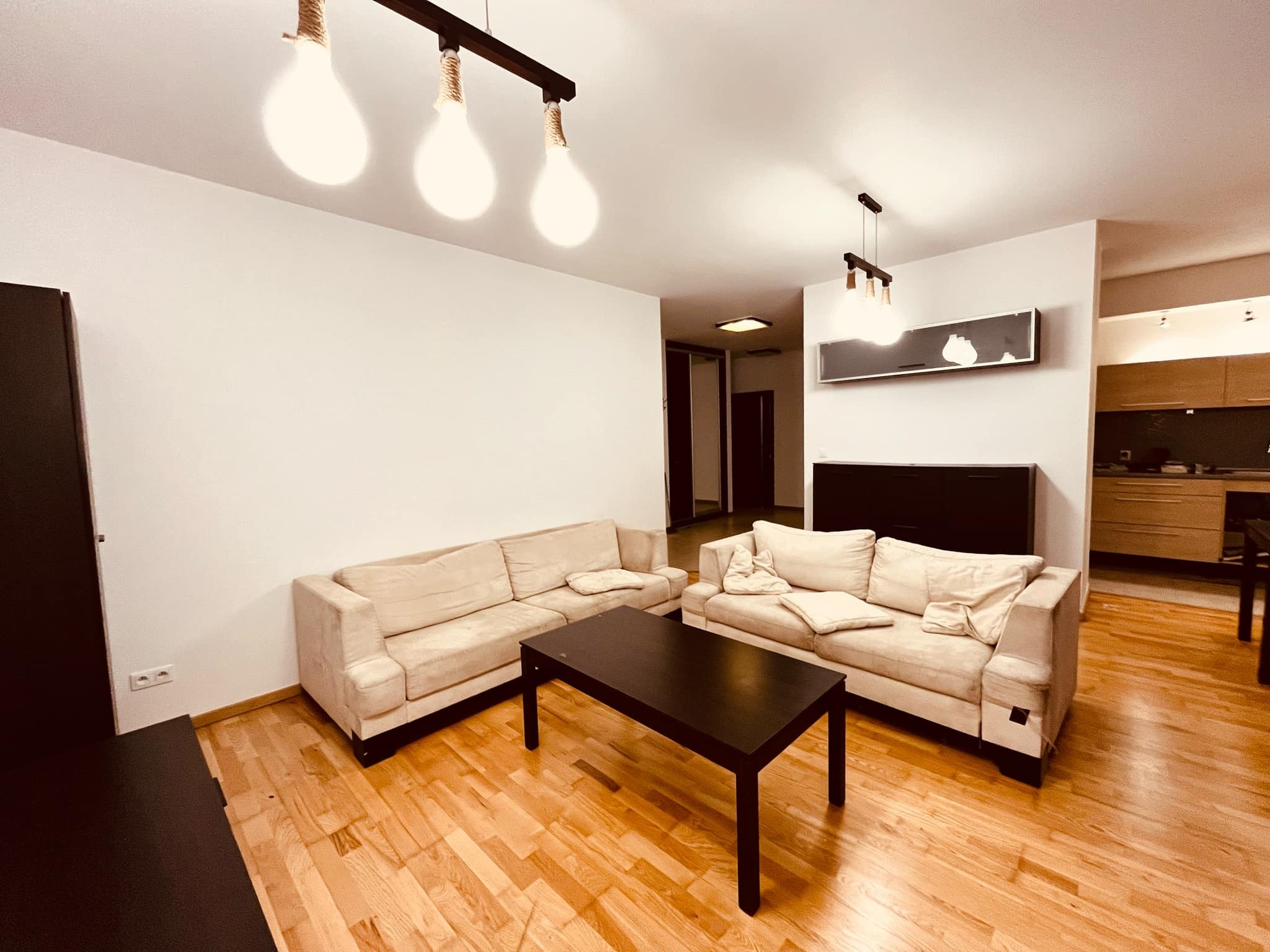 Jasne wnętrze pokoju dziennego po odświeżeniu ścian, z beżowymi sofami, ciemnym stolikiem kawowym i drewnianą podłogą, oświetlone lampami z dekoracyjnymi sznurami.