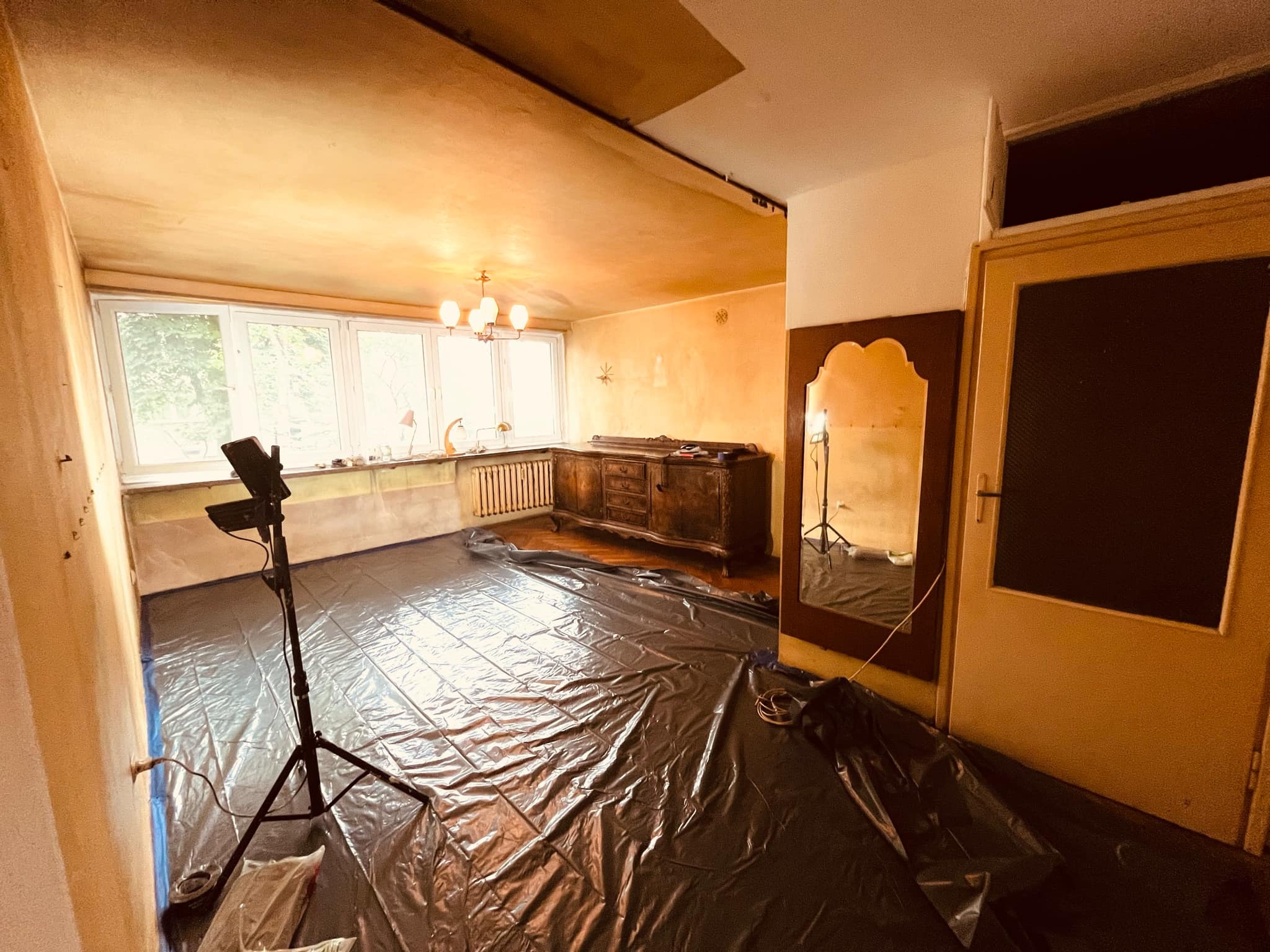 Pokój przygotowany do malowania: podłoga zabezpieczona czarną folią, widoczny statyw z lampą oświetleniową, ściany z widocznymi śladami po wcześniejszych pracach, stare meble przy ścianie.