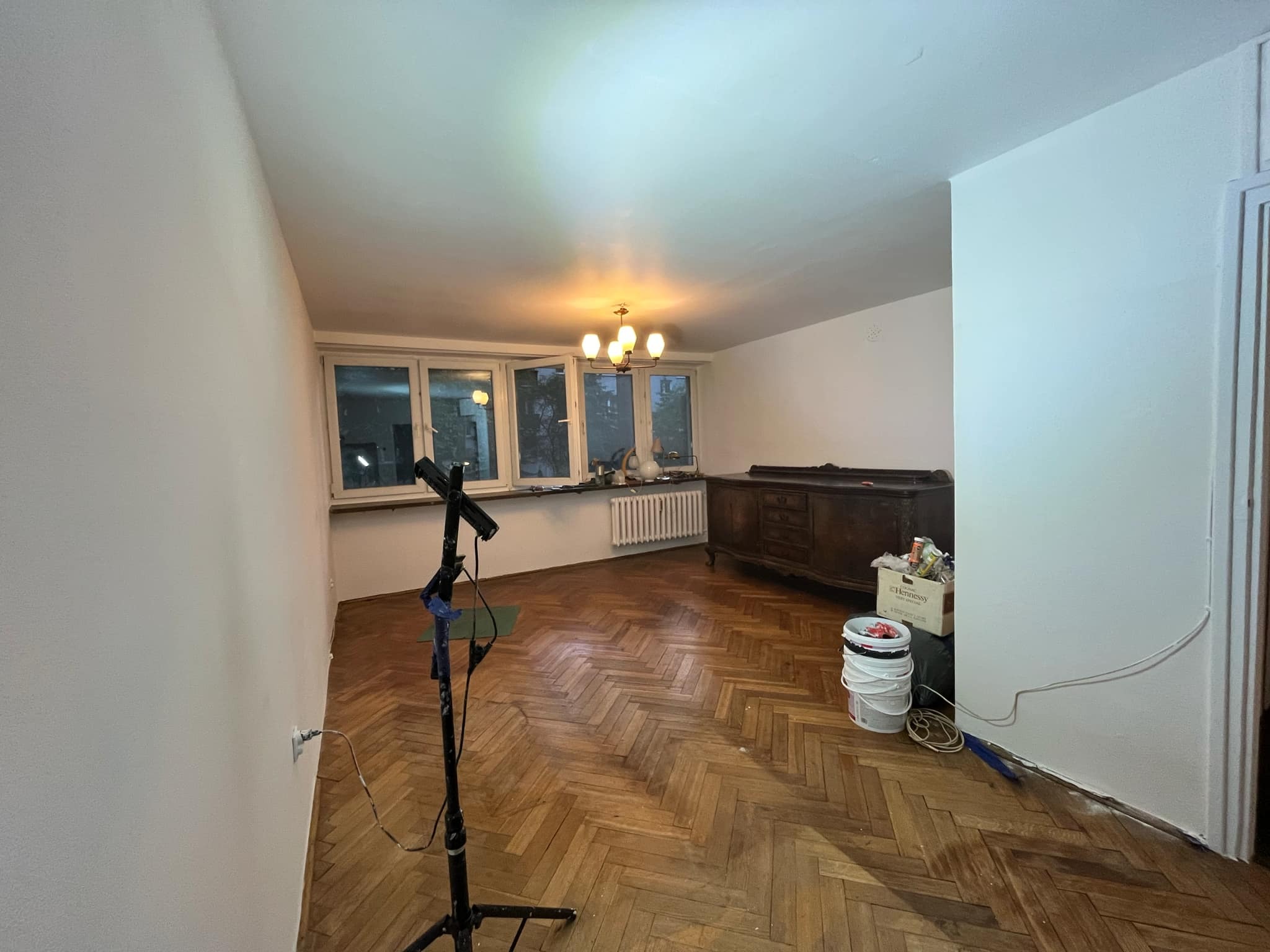 Pokój przed malowaniem: widoczny statyw oświetleniowy, stare meble, podłoga w jodełkę i ślady po taśmie malarskiej przy podłodze.