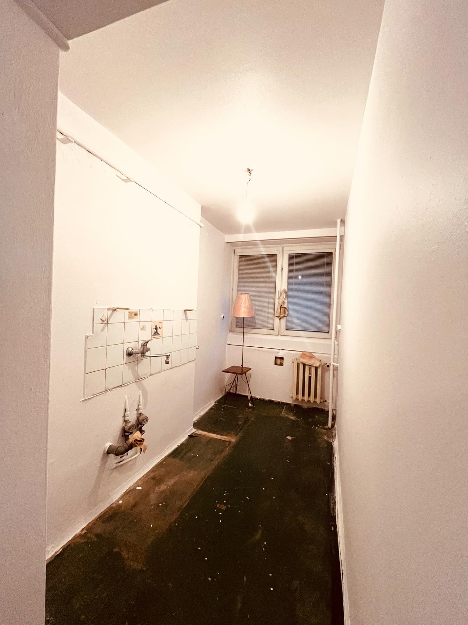 Wnętrze małej kuchni w trakcie remontu: ściany pomalowane na biało, częściowo usunięte płytki nad zlewem, odsłonięte rury, stara podłoga z resztkami wykładziny, grzejnik pod oknem, lampka na stoliku.