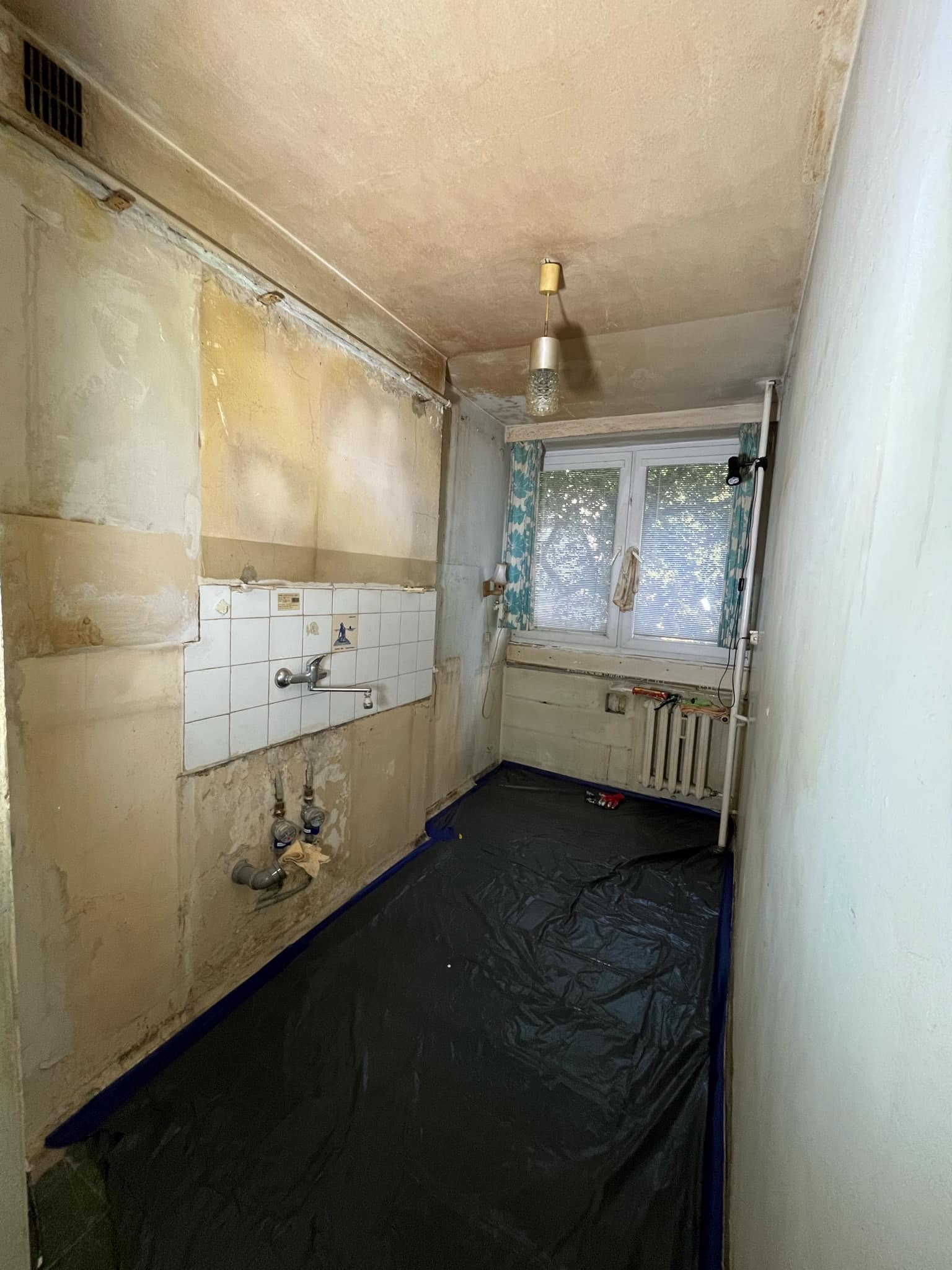 Przygotowanie do malowania kuchni: ściany z resztkami starej farby i płytek, zabezpieczona podłoga folią malarską, widoczne rury i zawory.