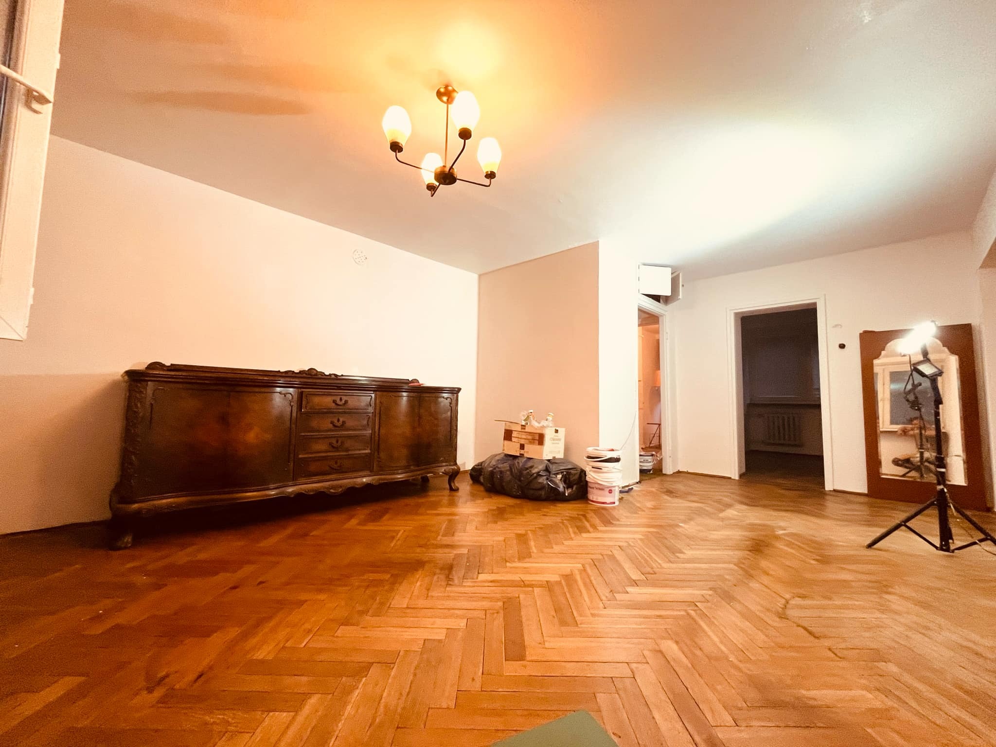 Przygotowania do malowania ścian w przestronnym pokoju w Lublinie, widoczny zabytkowy kredens odsunięty od ściany, zabezpieczona podłoga parkietowa w jodełkę, puszki z farbą i lampa oświetlająca...