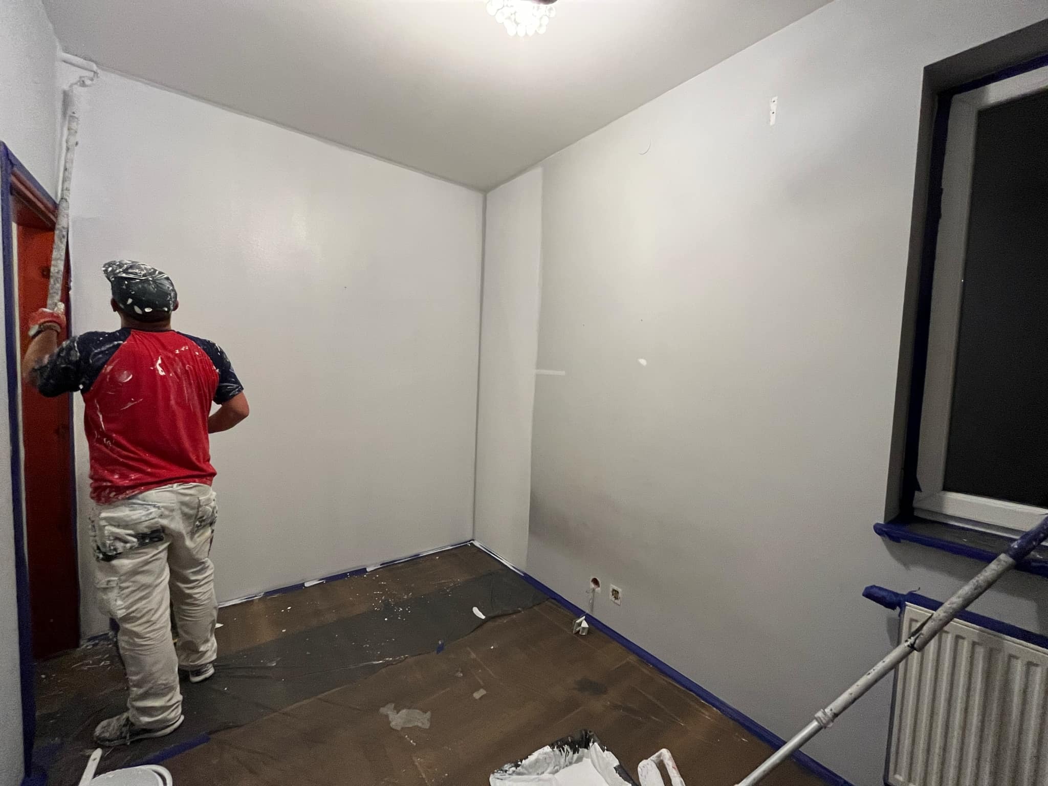 Malarz w czerwonej koszulce maluje ścianę na biało za pomocą wałka na długim kiju; widoczne zabezpieczenia taśmą malarską przy podłodze i futrynie drzwiowej.