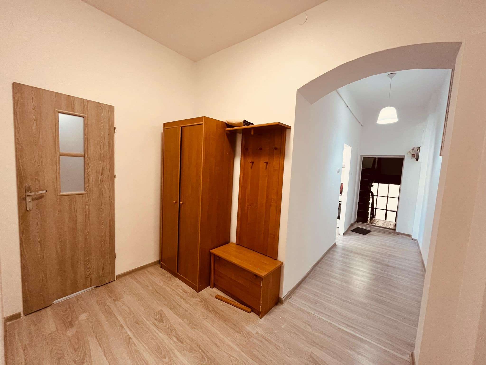 Jasne wnętrze przedpokoju z drewnianą podłogą, szafą, wieszakiem z siedziskiem i drzwiami z przeszkleniem.