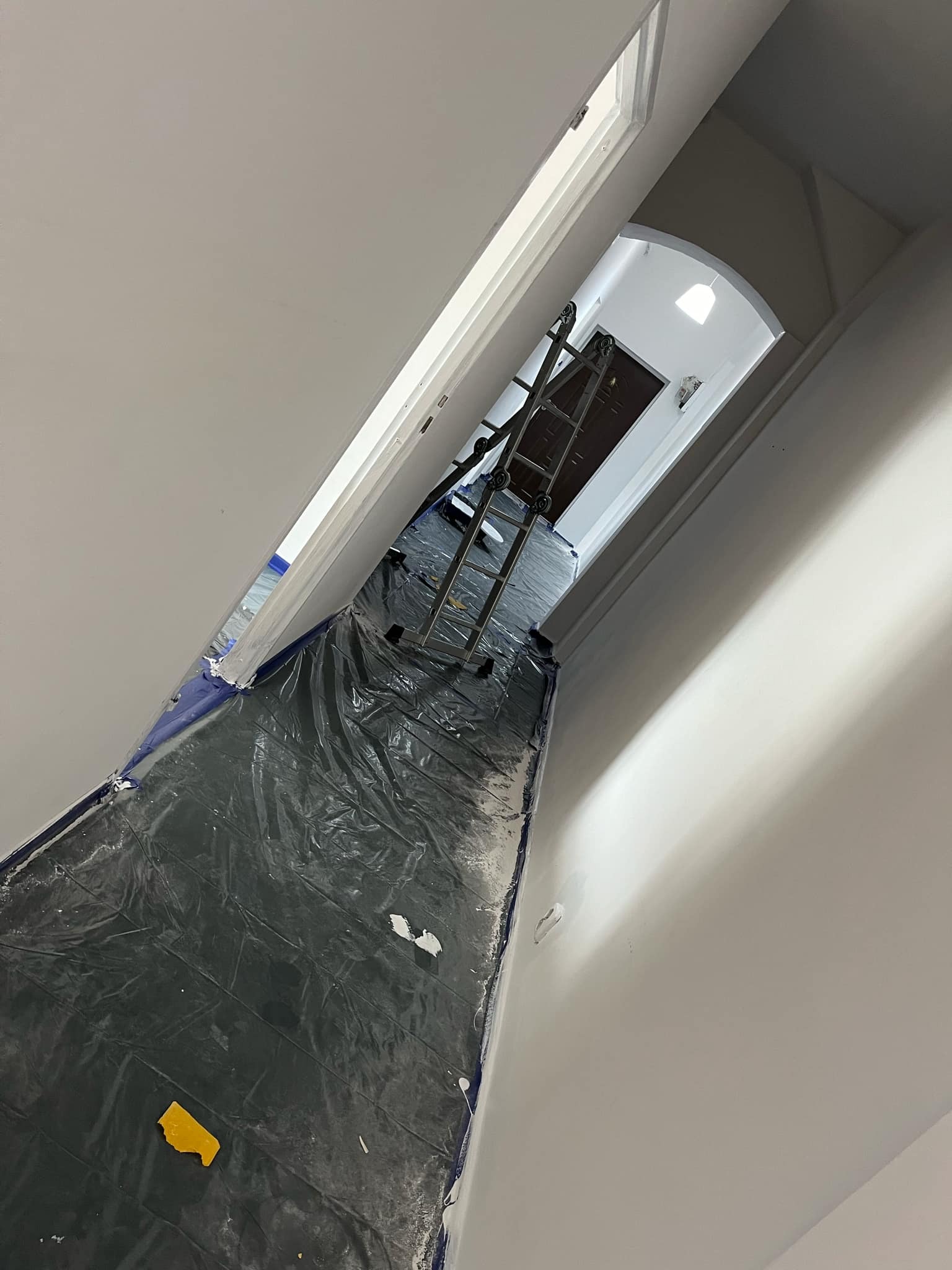 Korytarz w trakcie malowania: podłoga zabezpieczona folią, drabina rozstawiona w głębi, świeżo pomalowane ściany w odcieniach bieli.