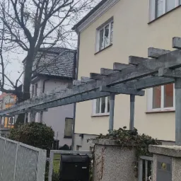 Metalowa pergola nad wejściem do domu z szarą bramą, czarną skrzynką pocztową i zielonym bluszczem na kamiennych słupkach w pochmurny dzień.