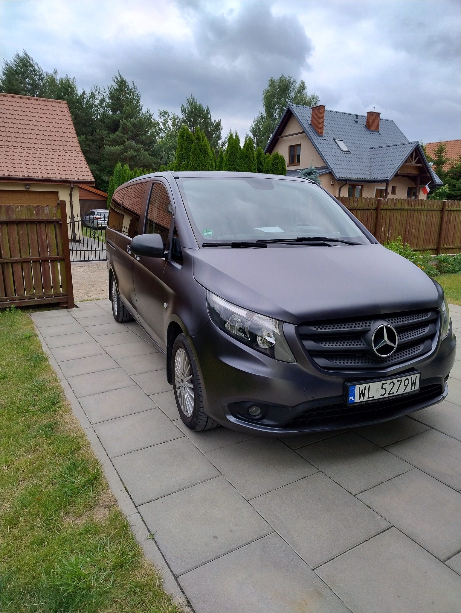 Szary Mercedes Vito, zaparkowany na posesji z kostki brukowej. W tle domy jednorodzinne i ogrodzenie. Tablica rejestracyjna z Warszawy.
