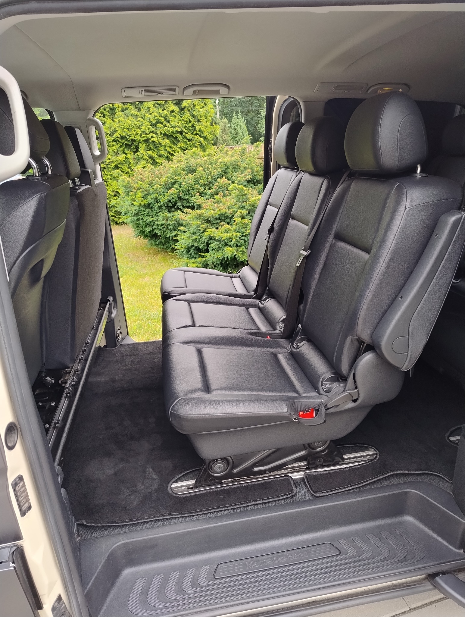 Wnętrze busa z trzema rzędami czarnych, skórzanych foteli. Widok na zielony krajobraz za oknem. Detal: szyny mocujące fotele i napis Mercedes-Benz na progu.