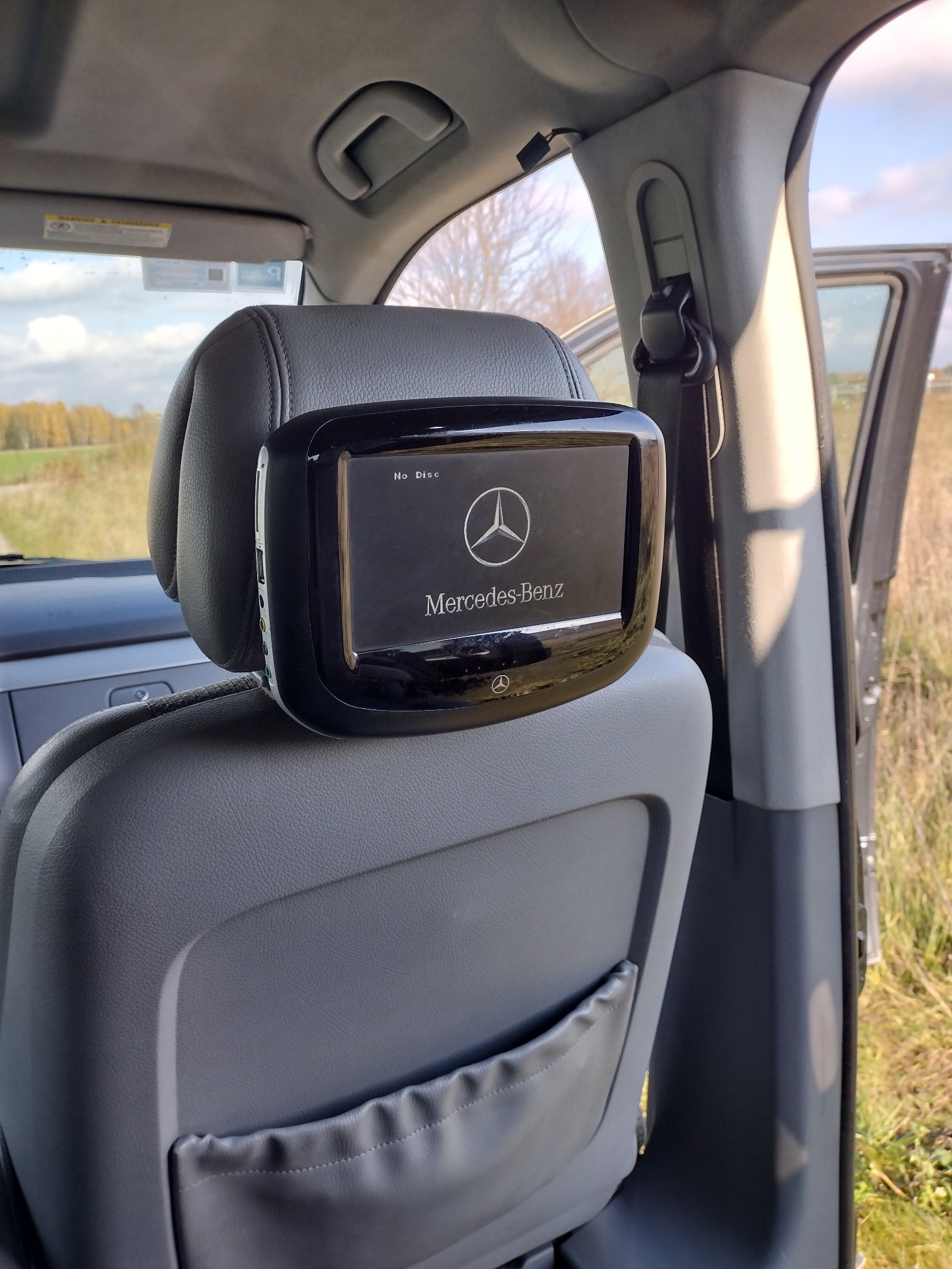 Wnętrze samochodu Mercedes-Benz w Serocku, z zamontowanym ekranem w zagłówku, wyświetlającym logo marki i komunikat 'No Disc', widok z tylnego siedzenia na otwarte drzwi i krajobraz za oknem.