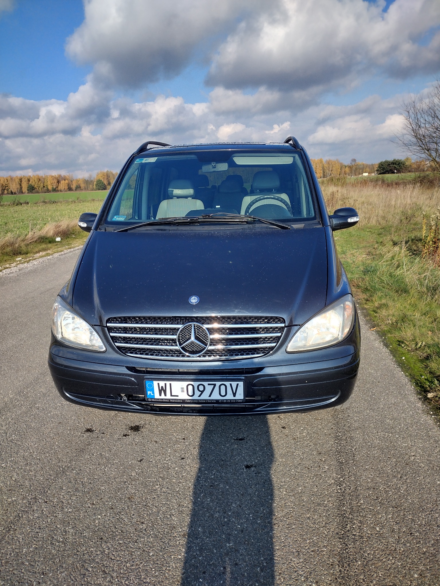 Czarny Mercedes Vito zaparkowany na asfaltowej drodze, widok z przodu, w tle jesienny krajobraz z polami i drzewami pod błękitnym niebem z chmurami.