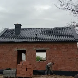 Nowy, czarny dach z kominem na surowym budynku z czerwonej cegły, pracownik kopie ziemię przy fundamencie, w tle widoczne pole i zachmurzone niebo.