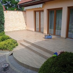 Bartłomiej Błaszkowski - Nowoczesny taras z płytek imitujących drewno, z niskimi schodami, otoczony zielenią, z widocznym fragmentem elewacji domu i narzędziami pracy.