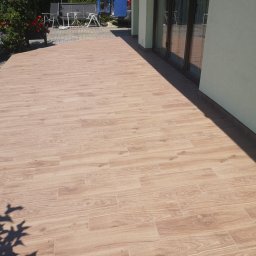 Bartłomiej Błaszkowski - Wyłożony płytkami taras w Sułkowicach, imitującymi drewno, z widocznymi drzwiami balkonowymi i fragmentem ogrodu w tle.