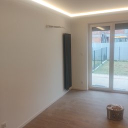 Bartłomiej Błaszkowski - Jasne wnętrze z drewnopodobną podłogą, grzejnikiem panelowym i oświetleniem LED podsufitowym, widok na ogród przez duże okno balkonowe, w tle zaparkowany żółty samochód.