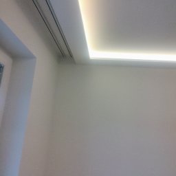 Bartłomiej Błaszkowski - Biały sufit z wbudowanym liniowym oświetleniem LED, widoczne kratki wentylacyjne w rogu pomieszczenia.