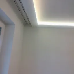 Biały sufit z wbudowanym liniowym oświetleniem LED, widoczne kratki wentylacyjne w rogu pomieszczenia.