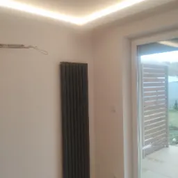 Szary, pionowy grzejnik panelowy na białej ścianie, z widoczną listwą LED pod sufitem i wyjściem na balkon z drewnianą balustradą.