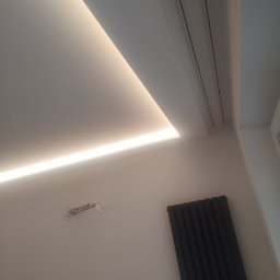 Bartłomiej Błaszkowski - Nowoczesny sufit z oświetleniem LED wzdłuż krawędzi, widoczny grzejnik panelowy na ścianie i wystające kable ze ściany.