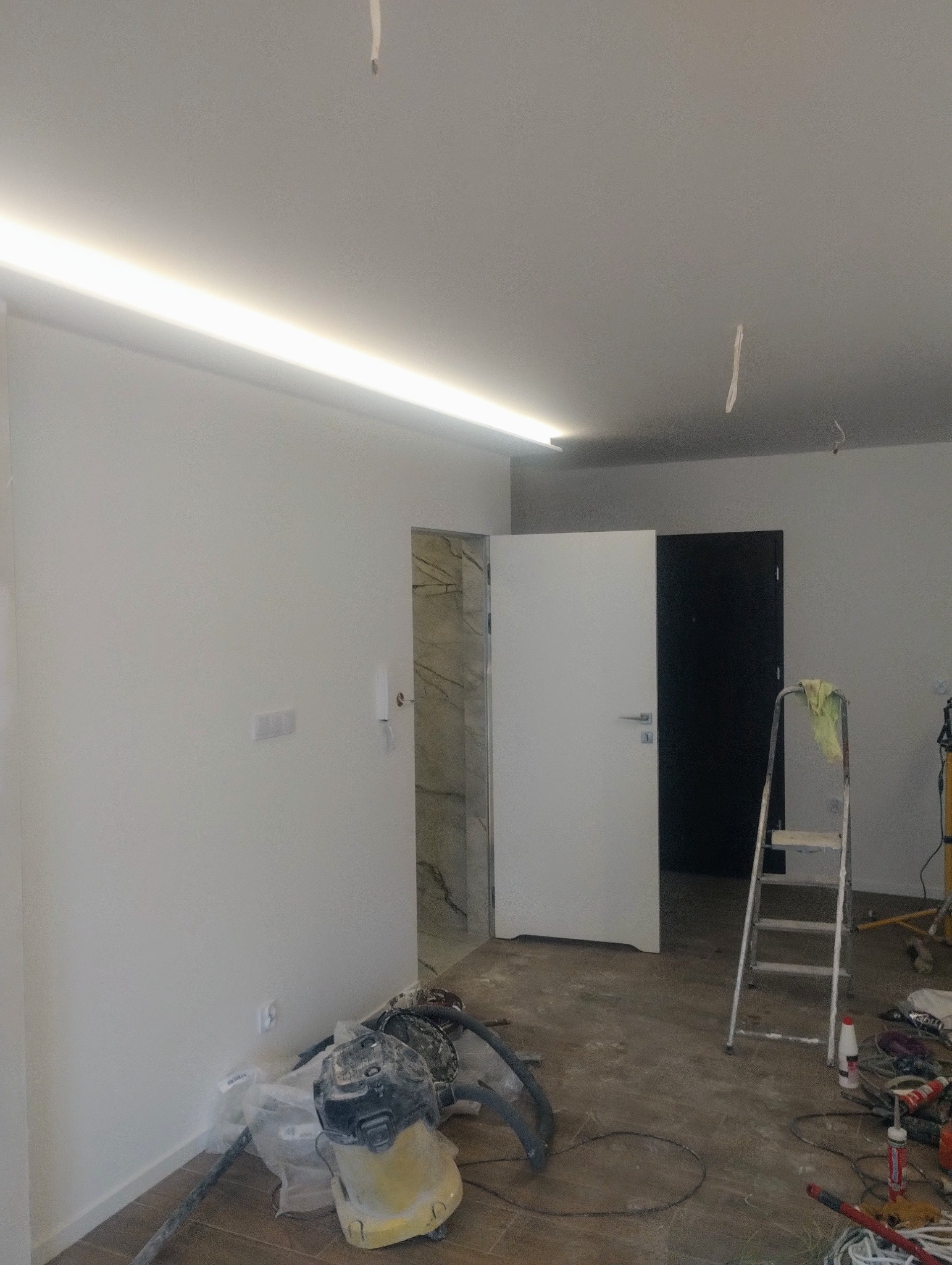 Wnętrze w trakcie prac wykończeniowych: zamontowane oświetlenie LED podsufitowe, widoczna łazienka z marmurowymi płytkami, drzwi wewnętrzne, drabina i odkurzacz przemysłowy.