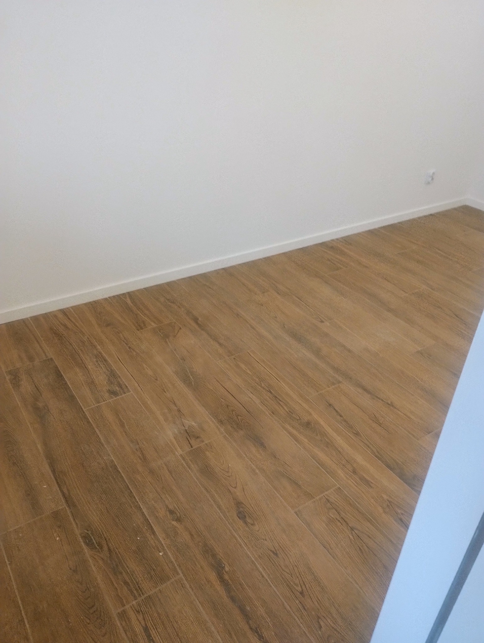 Ułożona pod kątem podłoga z płytek imitujących drewno w jasnym pomieszczeniu z białymi ścianami i listwami przypodłogowymi.