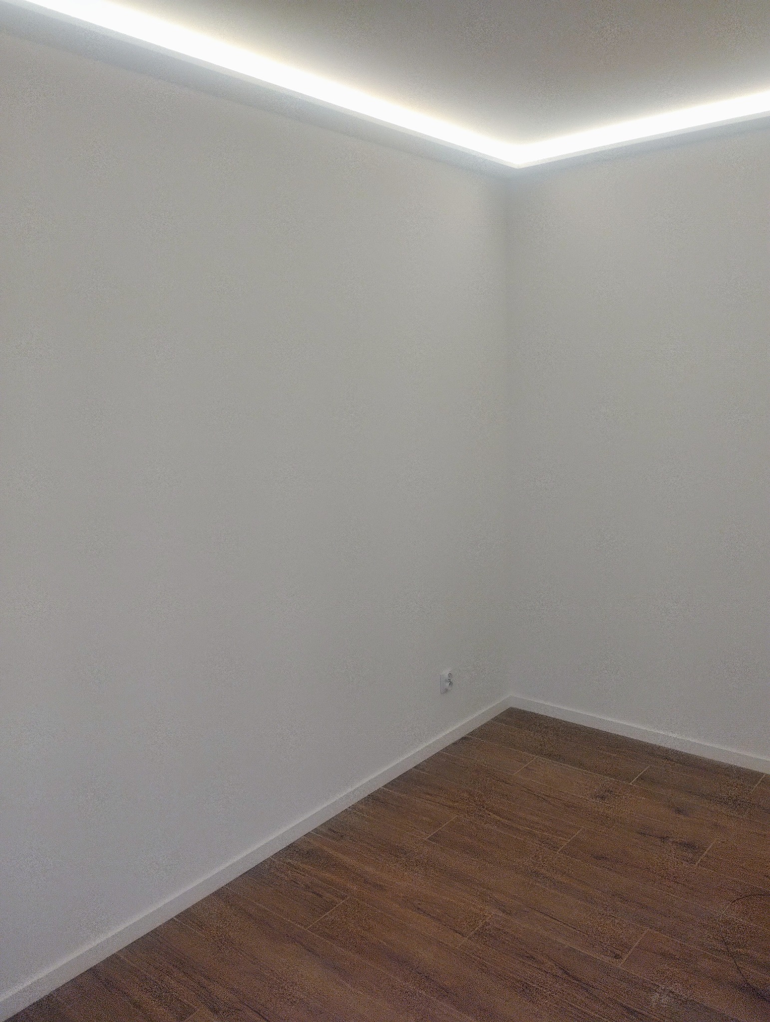 Pusty pokój po remoncie z drewnopodobną podłogą z płytek i oświetleniem LED wzdłuż sufitu, widoczne gniazdko elektryczne.