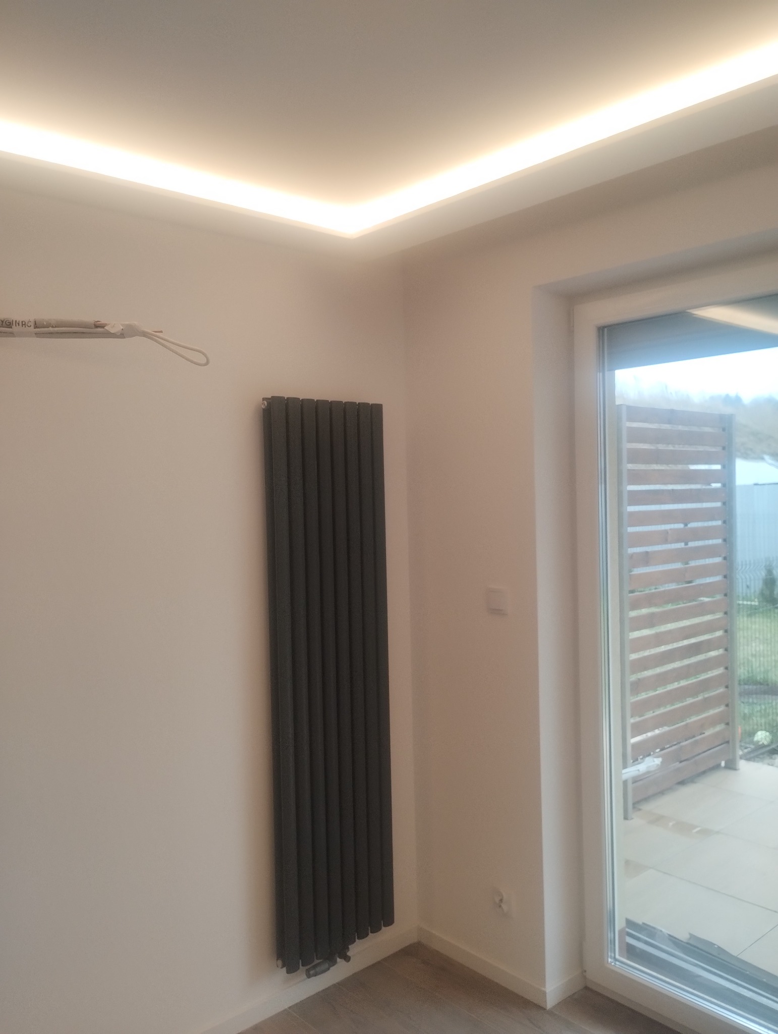 Szary, pionowy grzejnik panelowy na białej ścianie, z widoczną listwą LED pod sufitem i wyjściem na balkon z drewnianą balustradą.