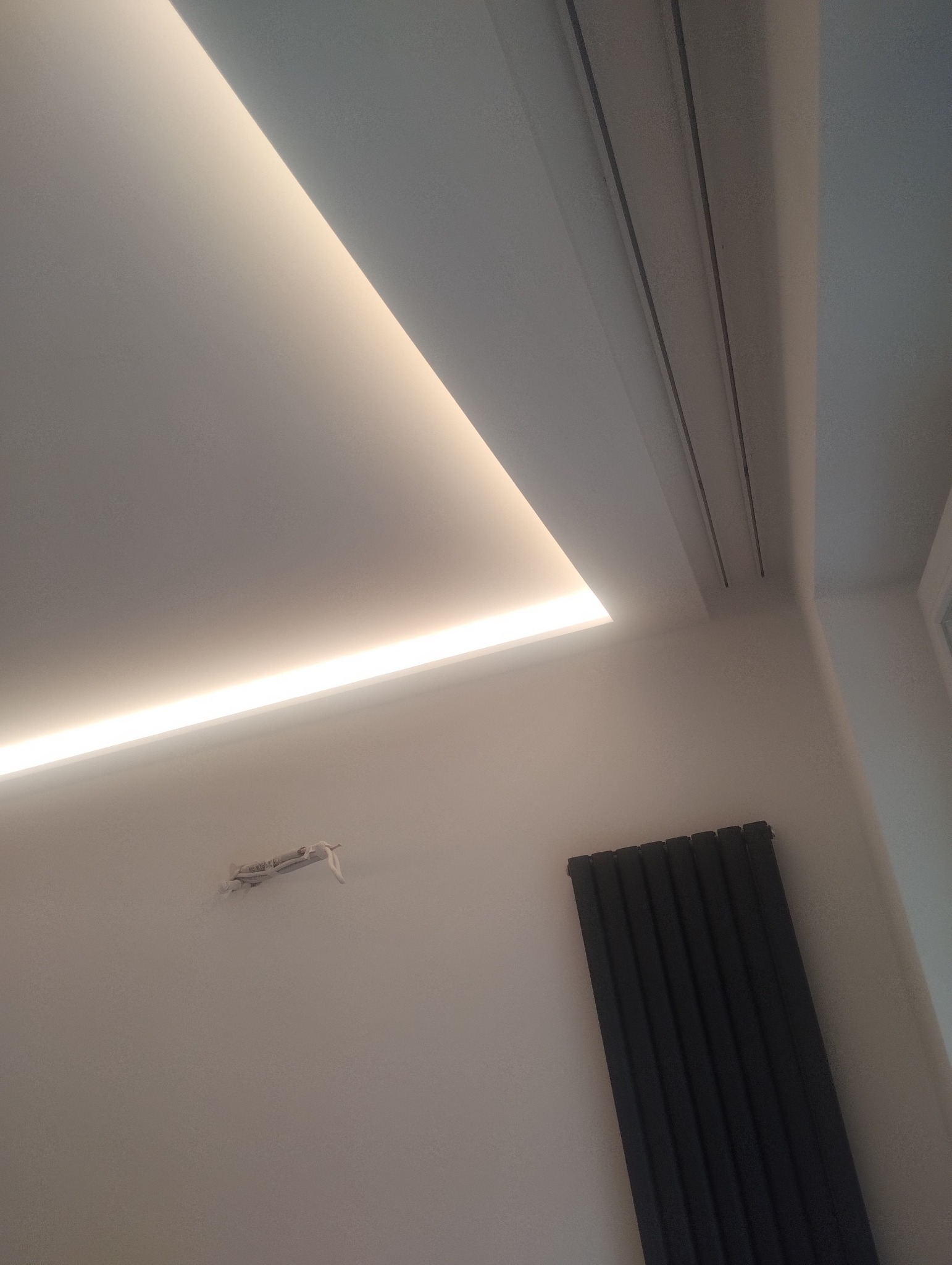 Nowoczesny sufit z oświetleniem LED wzdłuż krawędzi, widoczny grzejnik panelowy na ścianie i wystające kable ze ściany.