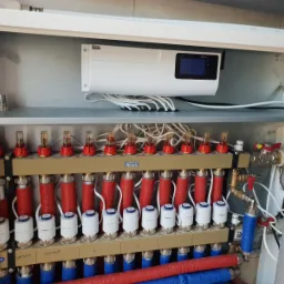 Rozdzielacz ogrzewania podłogowego w szafce, z widocznymi rurami zasilającymi i powrotnymi, zaworami termostatycznymi, przepływomierzami oraz sterownikiem elektronicznym TECH.