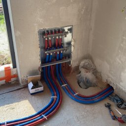 Modern System - Rozdzielacz ogrzewania podłogowego z czerwono-niebieskimi rurami w surowym stanie, widoczne narzędzia i karton po akcesoriach Cluro